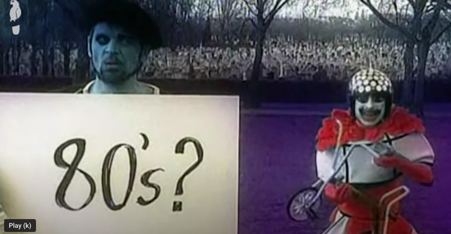 A man with blue skin wearing a beret holds up a placard with "80's?" written on it while some kind of goblinoid creature (who is actually David Walliams would you believe it?) crouches in the background grinning unnervingly and holding up a child's tricycle.
